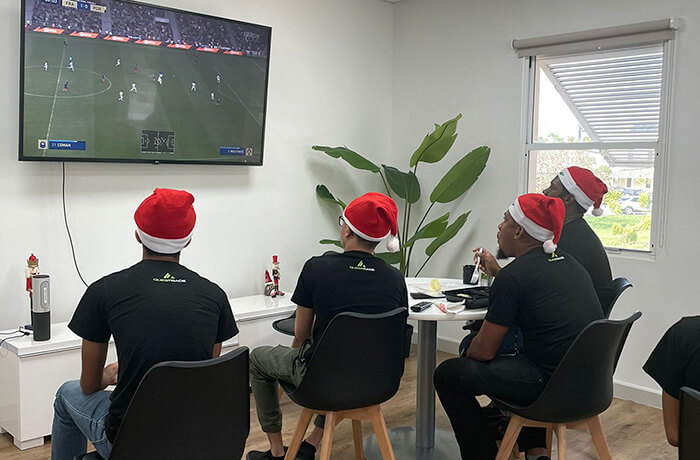 Four people wering a christmas hat and watching a soccer game on the TV