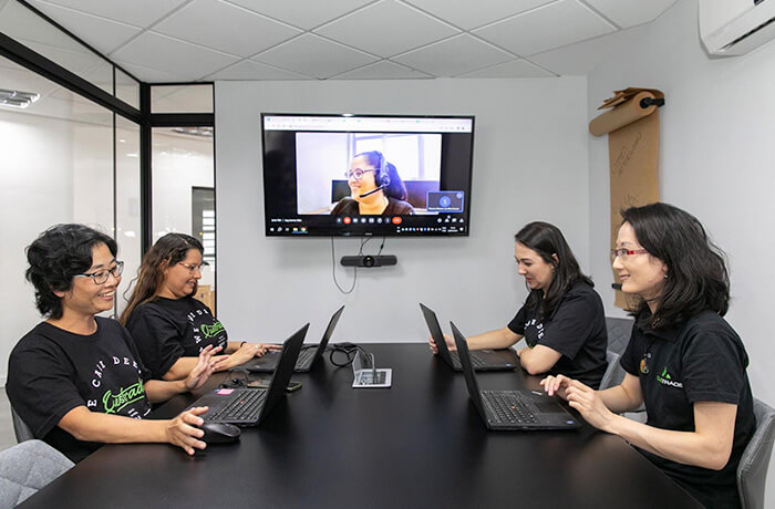 Four women sitting in a meeting room and one woman joining the meeting virtually